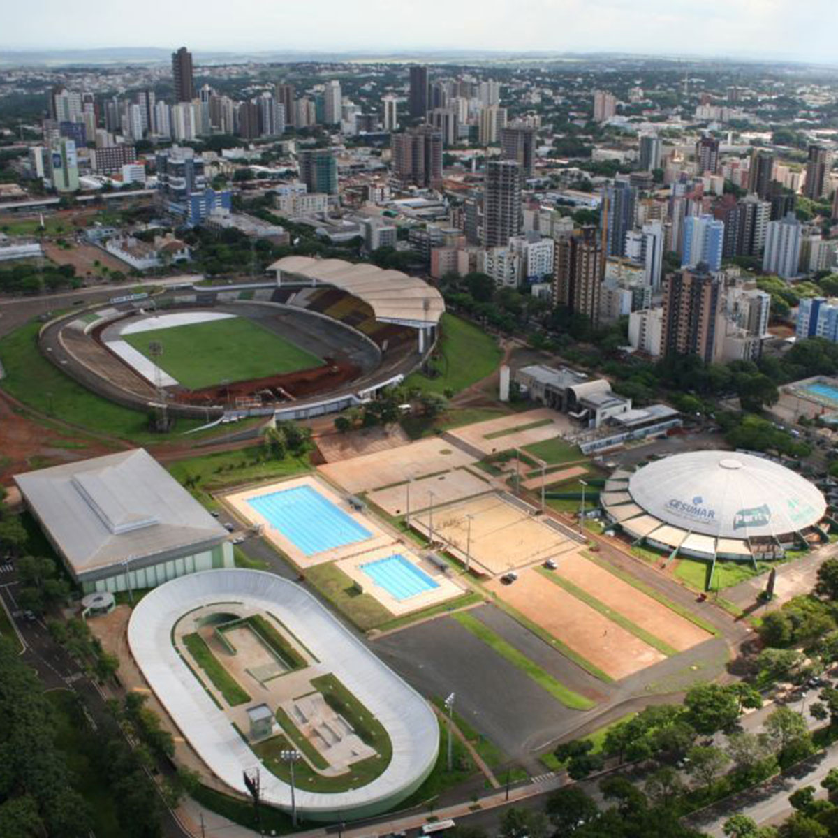 Vista aérea da Vila Olímpica de Maringá, com pistas, piscinas e o horizonte da cidade ao fundo.